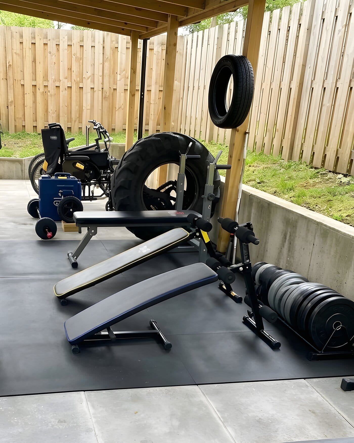 Three weight benches, a rack of plates, dumbbells and a tractor tyre laid out on rubber matting under a wooden pergola